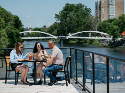 three people sitting on a patio table overlooking a river, a pedestrian crossing bridge in the background