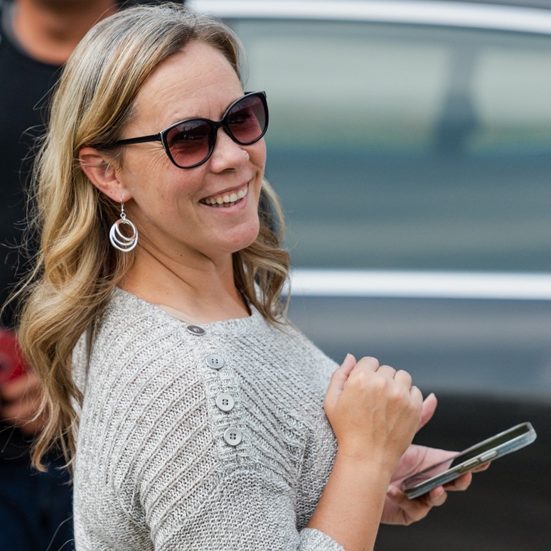 A woman wearing sunglasses and a sweater smiles in front of a car.