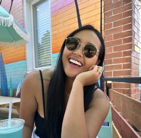 a woman with long hair and sunglasses smiling at a blue picnic table with a blue smoothie beside her