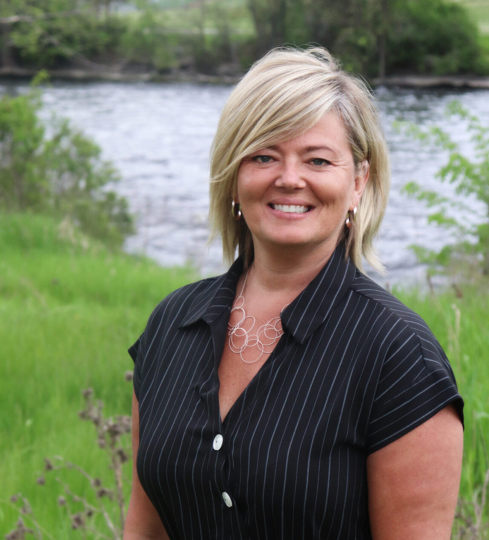 a woman posing for a headshot with blonde hair, wearing a black dress, out in nature by a river