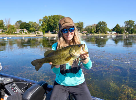 ashley (white woman with long blonde hair) kneeling on a fishing boat showing off the bass she caught