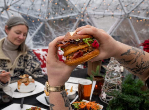 a hand holding up a burger while dining in a snowglobe tent outside in the winter