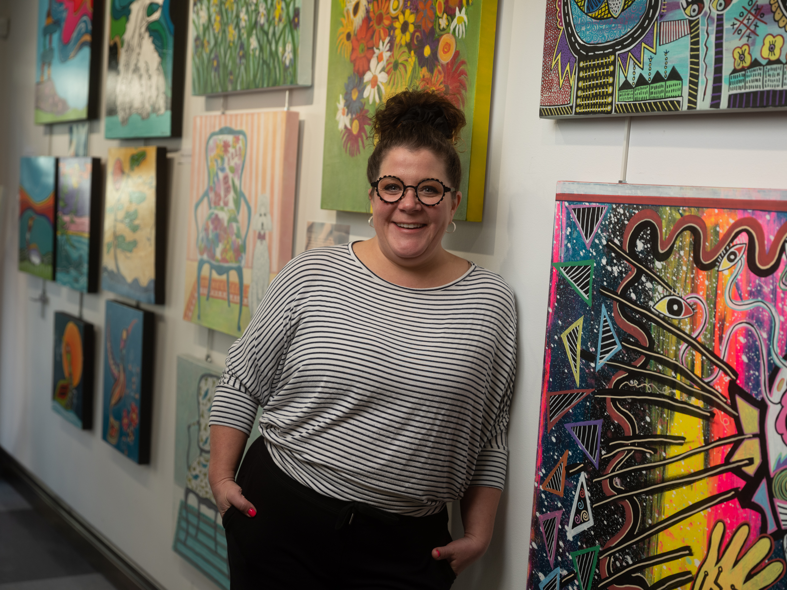 a woman with light skin, dark curly hair and a striped shirt standing against the wall of an art gallery