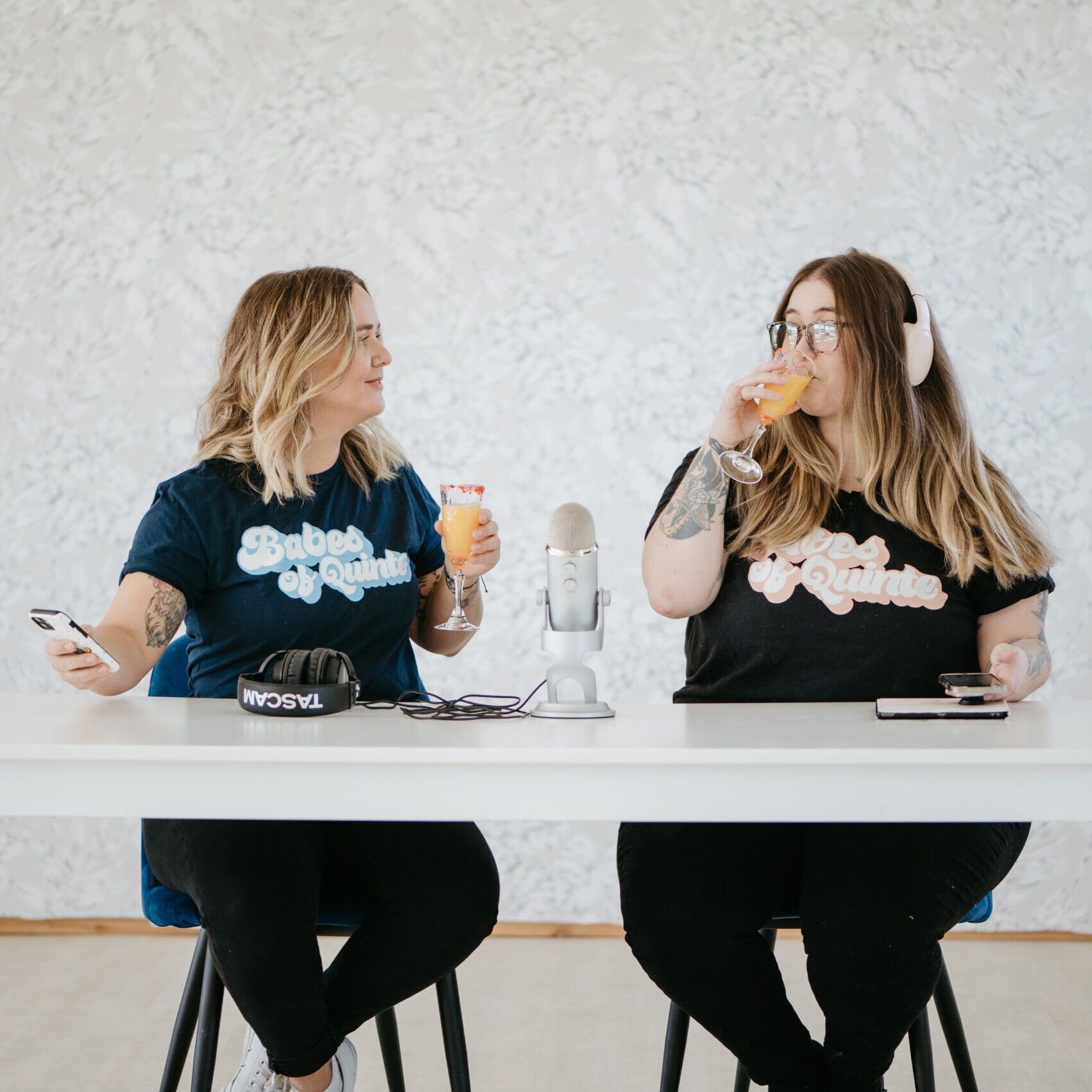 two women (light skin, blonde hair) sitting at a table drinking mimosa's, on their cell phones with a podcast recording microphone in the middle