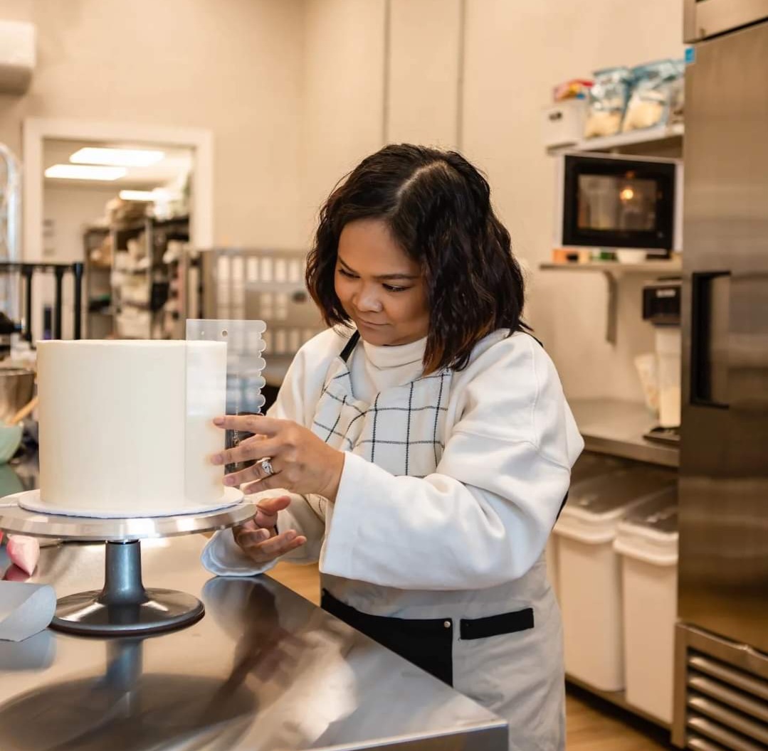 a woman (medium skin, dark brown short hair) putting icing on a cake in a professional bakery