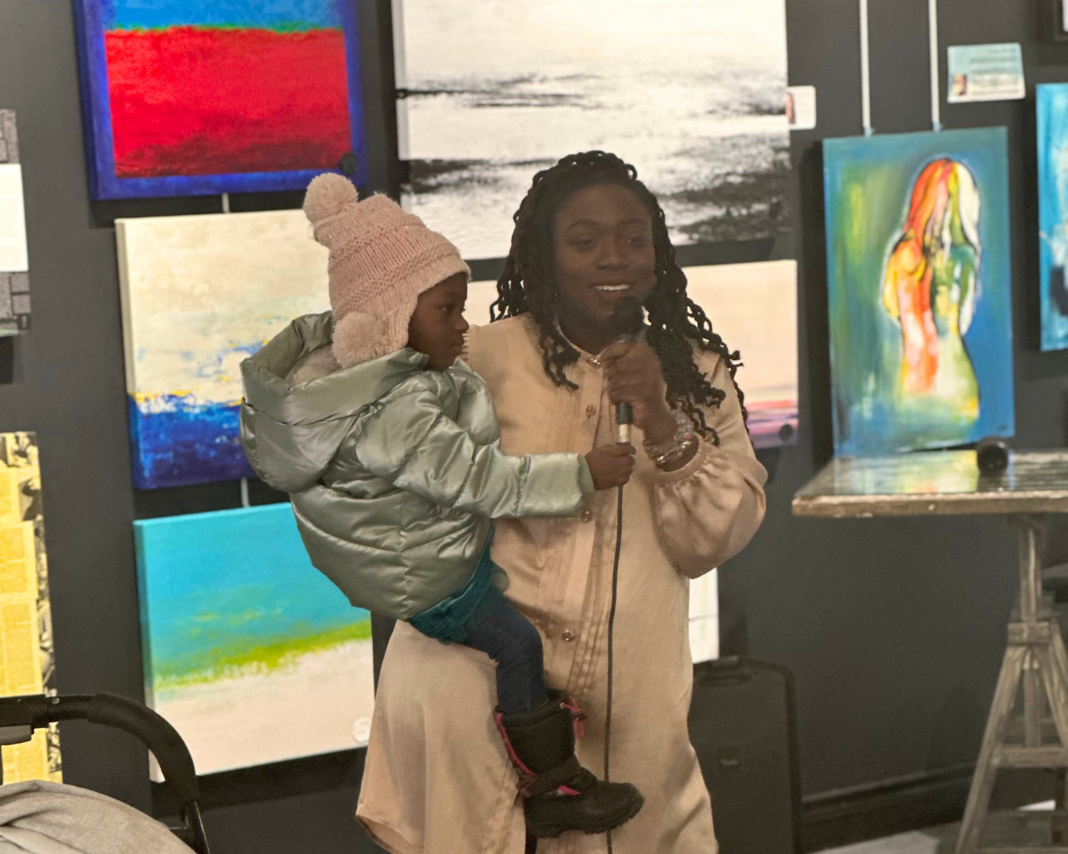 a woman speaking with a microphone while holding a small child, standing in an art gallery presenting to an audience