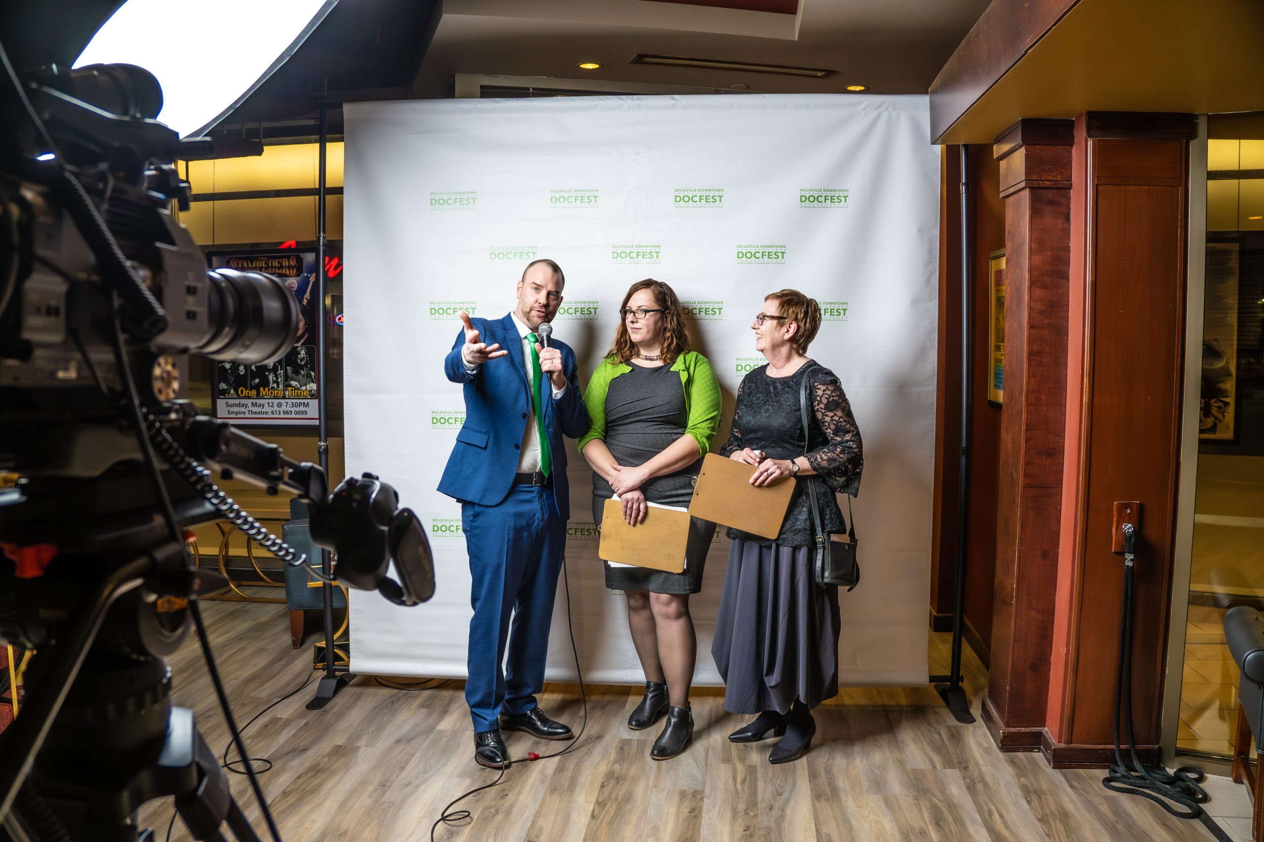 a man interviewing two women in front of a white and green DocFest back drop with a camera recording them