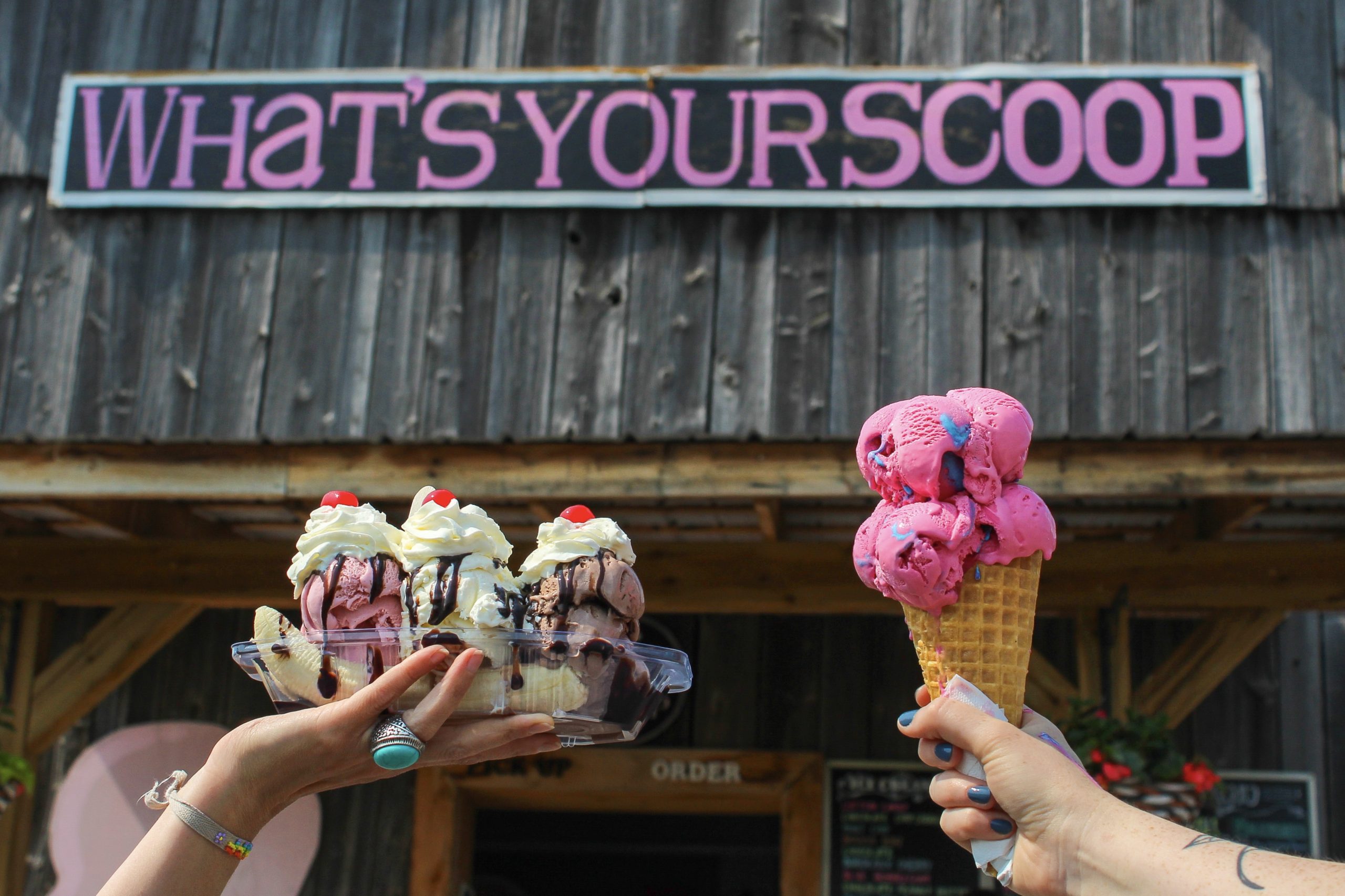 two hands holding up ice cream in front of the what's your scoop sign, one hand has a big cone of pink cotton candy ice cream and the other is holding a banana split