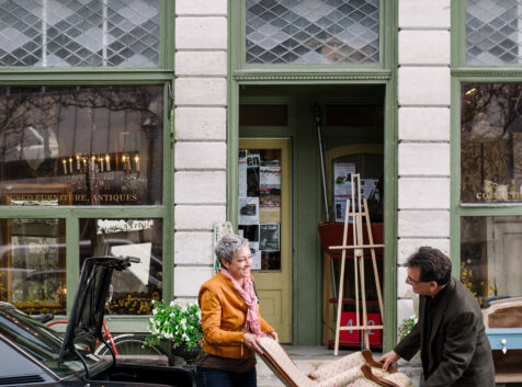 Image of two people trying to put an antique chair into the trunk of their car parked in front of a vintage shop